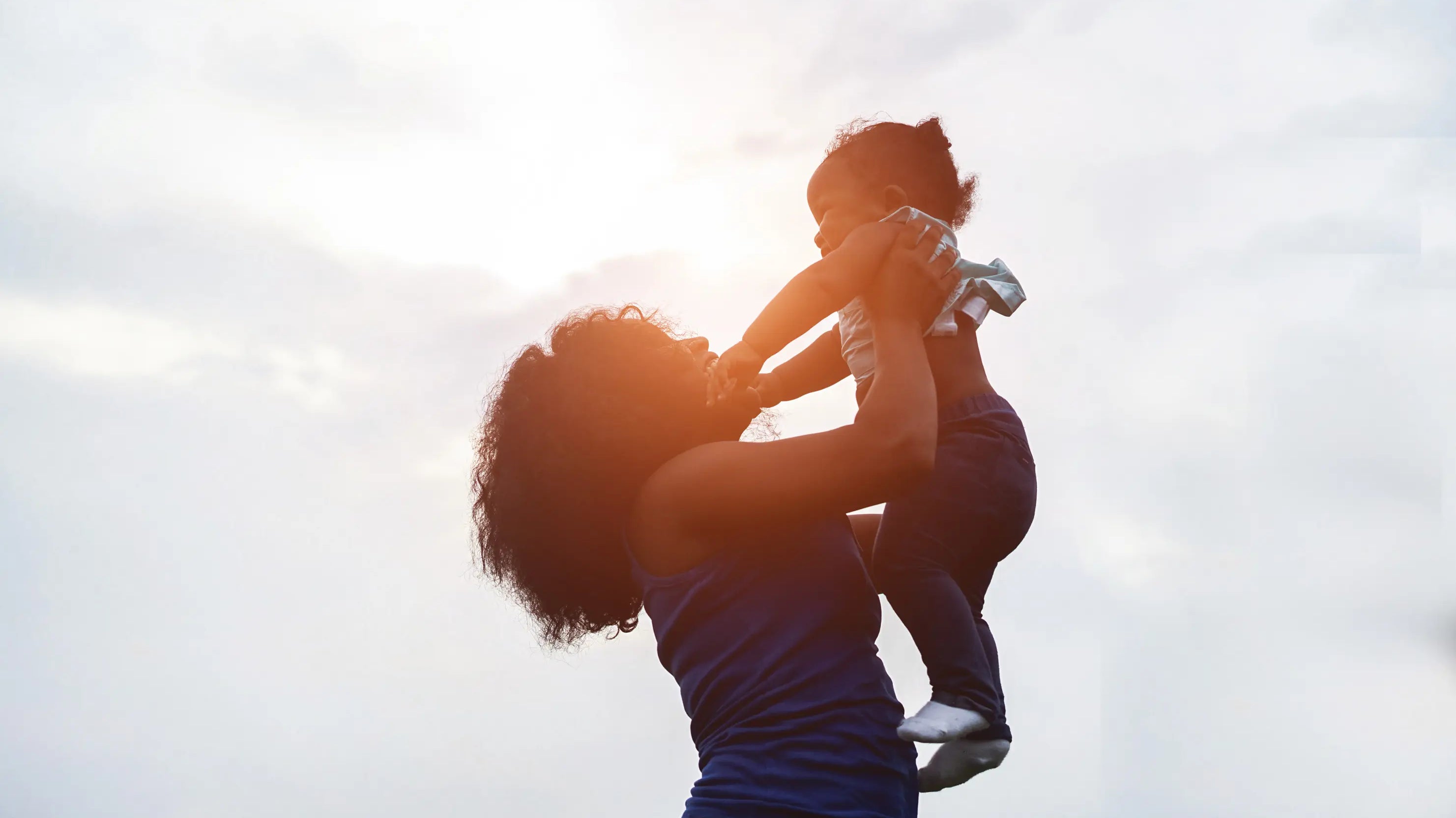 Mother holding a young child up in the air outdoors at sunset, both smiling and facing each other, with a soft glowing sky in the background.