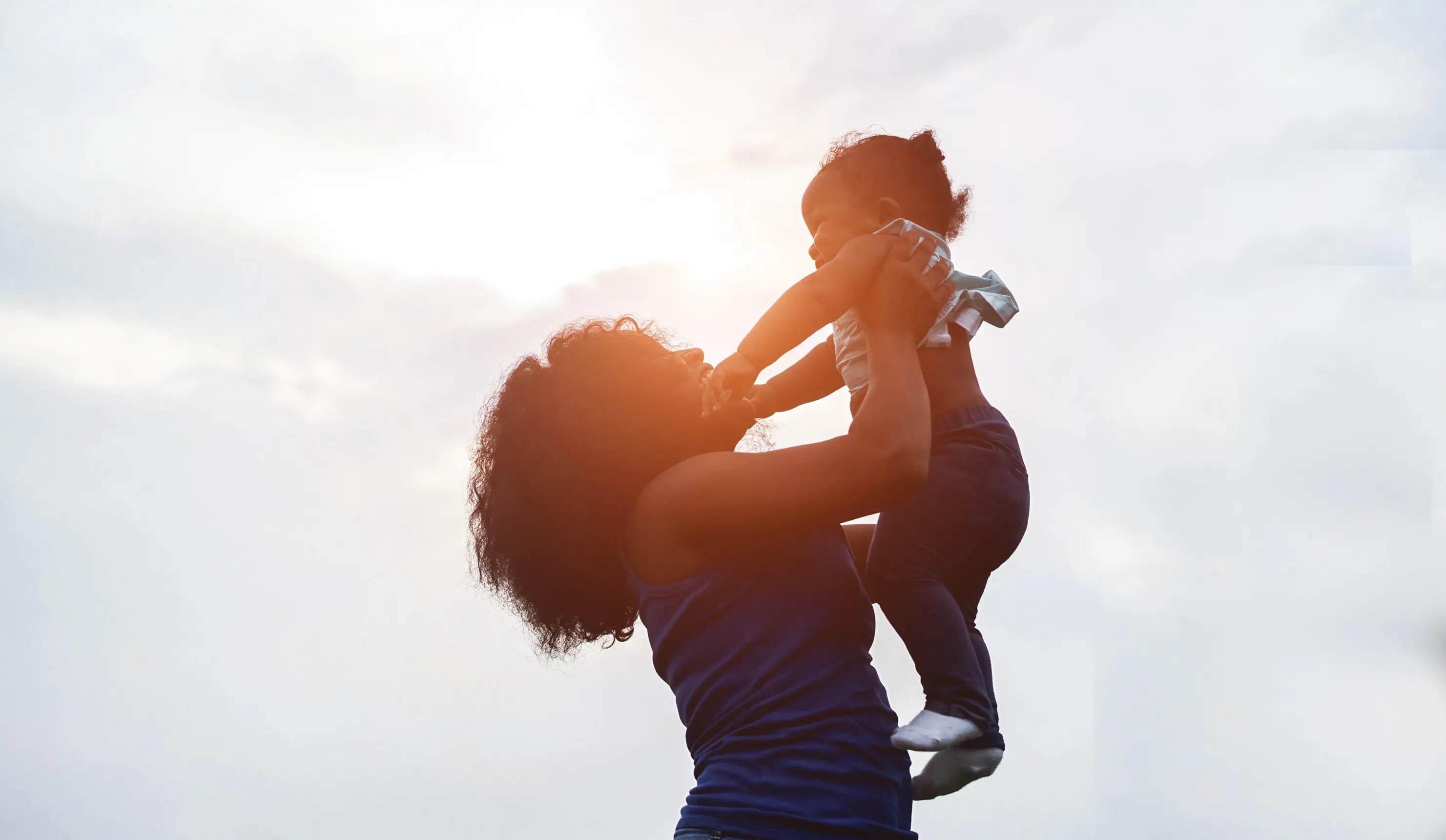 Mother holding a young child up in the air outdoors at sunset, both smiling and facing each other, with a soft glowing sky in the background.